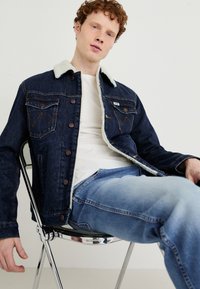 Denim jakna s krznim ovratnikom, z metalnimi gumbi, žepoma na prsih in klasičnim krojem. Svetlo modri kavbojke dopolnjujejo oblačilo.
