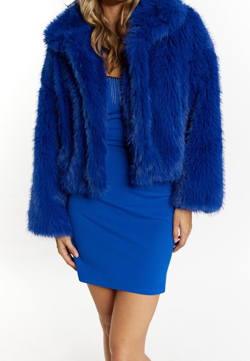 Femme portant une veste en fausse fourrure bleu vif sur une robe bleu ajustée sans manches, montrée du cou jusqu'à mi-cuisse sur un fond blanc.