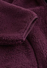 Fleece-stof i dyb lilla med en blød, tekstureret overflade. Har en glat kant langs kanterne, som forbedrer holdbarheden og strukturen.