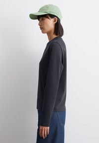 Sort langærmet top med rund neckline, parret med en grøn kasket. Stoffet fremstår glat og bomuldsagtigt med minimal detaljering.