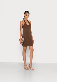 Vestido castanho justo com decote arredondado e alças de halter, complementado por uma clutch decorativa e sandálias de salto bege.