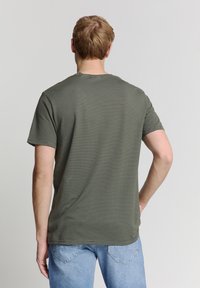 Olijfgroene gestreepte t-shirt met een ronde hals en korte mouwen, weergegeven op een persoon van achteren, gecombineerd met lichtblauwe spijkerbroeken.