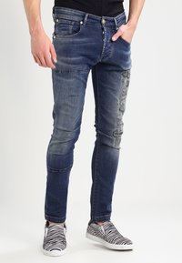 Persona che indossa jeans slim-fit blu sbiaditi con dettagli strappati e scarpe slip-on bianco e nere con stampe zebrate e leopardate.