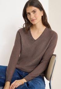 Brązowy sweter dzianinowy z dekoltem w serek, długimi rękawami, ściągaczami przy nadgarstkach i miękką fakturą. W połączeniu z niebieskimi dżinsami w pozycji siedzącej.