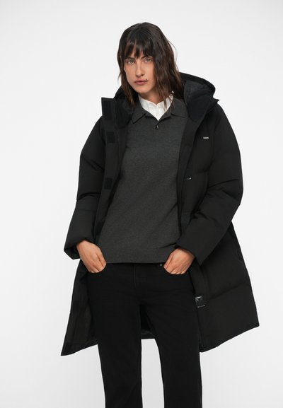 Čierny páperový kabát s kapucňou, s hladkým, polstrovaným dizajnom. Modelka na sebe má šedý sveter a čierne nohavice, stojí na bielom pozadí.