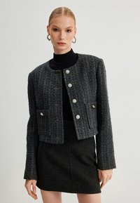 Grijze scheerjack met een cropped ontwerp, featuring metalen knopen, lange mouwen en voorzakken; gecombineerd met een getailleerde zwarte rok.