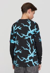 Nastolatka w czarnej i niebieskiej koszulce z krótkim rękawem w wzór błyskawicy, założonej na czarną koszulę z długim rękawem, w połączeniu z szarymi dżinsami.