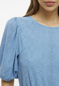 Ljusblå blus med texturerad tyg med intrikata mönster med utskärningar och korta puffärmar. Rund halsringning, slät finish.