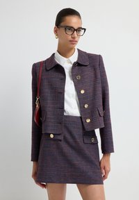 Completo giacca in tweed e mini gonna di colore blu scuro con pagliuzze rosse. Presenta bottoni dorati, un colletto e tasche. Camicia bianca sotto.