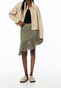 Beige spijkerjack, zwart cropped topje, olijfgroene gebreide asymmetrische rok met gerafelde zoom, en tan slip-on schoenen met zwarte zolen.