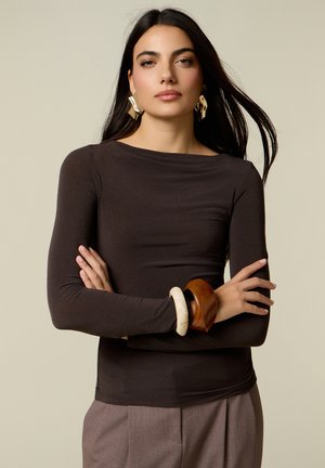 Mujer con cabello largo y oscuro que lleva una blusa ajustada de manga larga de color marrón oscuro, pantalones topo, grandes pendientes dorados y pulseras gruesas de madera y marfil.