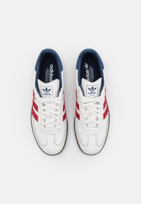White leather sneakers with red suede stripes, a blue tongue, and beige rubber outsole. Features round laces and a padded collar.