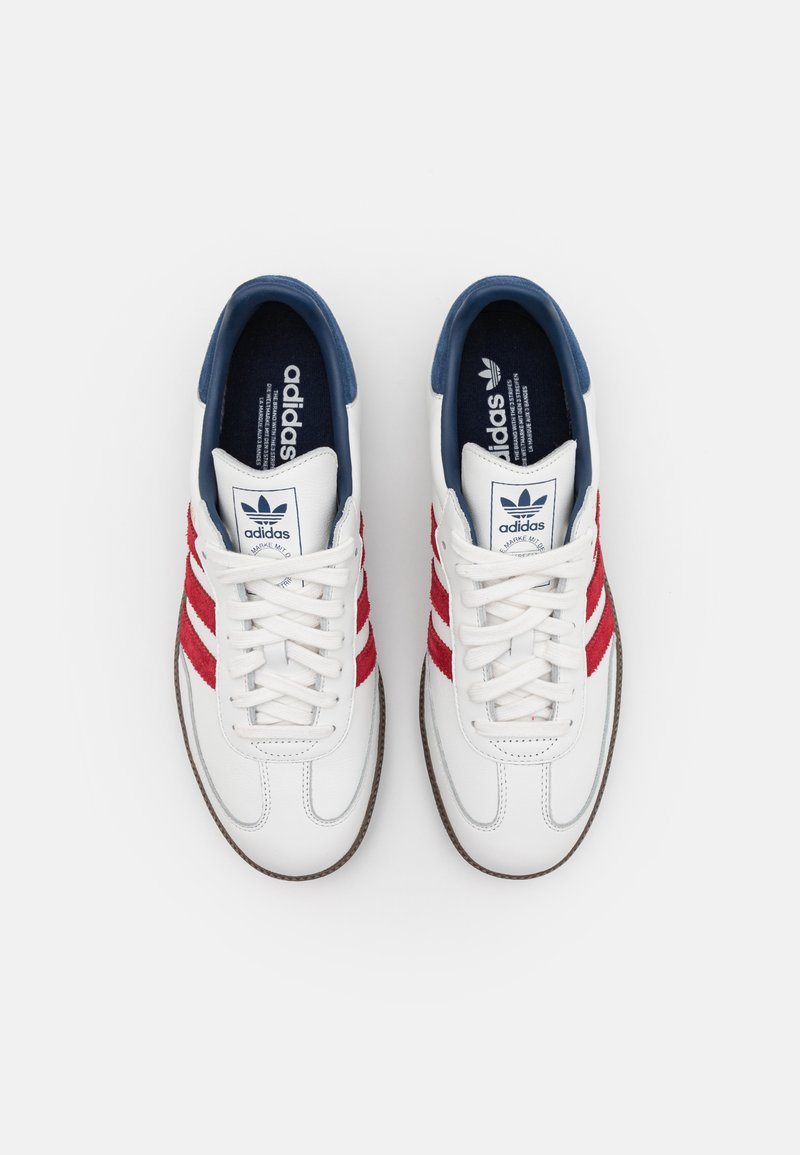 Zapatillas de cuero blanco con rayas de ante rojo, una lengüeta azul y suela de goma beige. Presentan cordones redondos y un cuello acolchado.