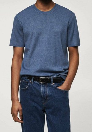 Man draagt een effen blauw shirt met korte mouwen, in een medium gewassen spijkerbroek met een zwarte riem, één hand in de zak tegen een effen achtergrond.