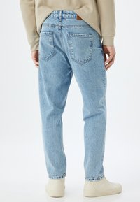 Jeans in denim azzurro chiaro con una vestibilità rilassata, due tasche posteriori e dettagli cuciti. Indossati con scarpe beige e un top abbinato.