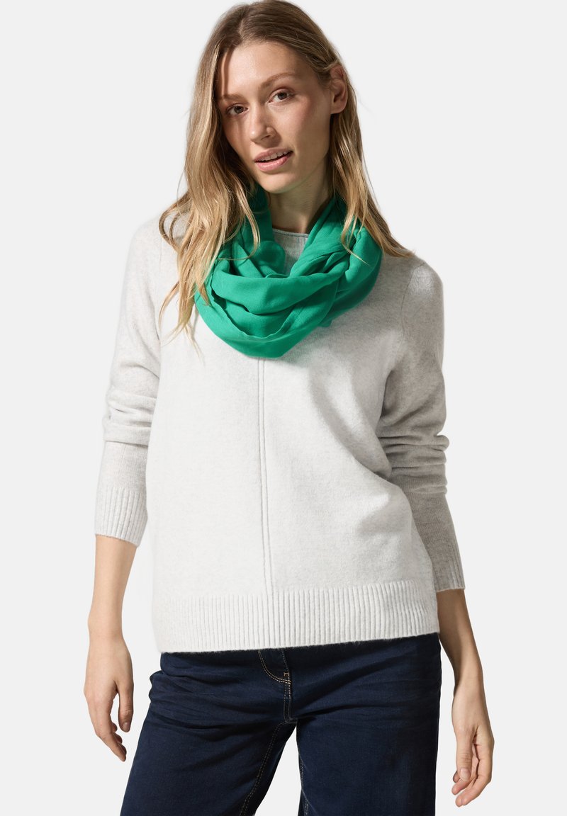 Grüner Infinity-Schal aus leichtem Stoff, getragen über einem hellgrauen Pullover mit strukturiertem Rippensaum. Dunkle Jeans runden das Outfit ab.