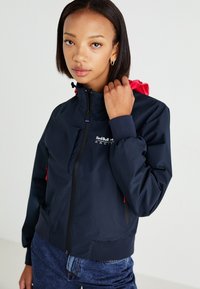 Chaqueta cortavientos de color marino con cuello alto, cremallera negra, cordón rosa y puños elásticos. Logo de "Red Bull Racing" en el pecho.