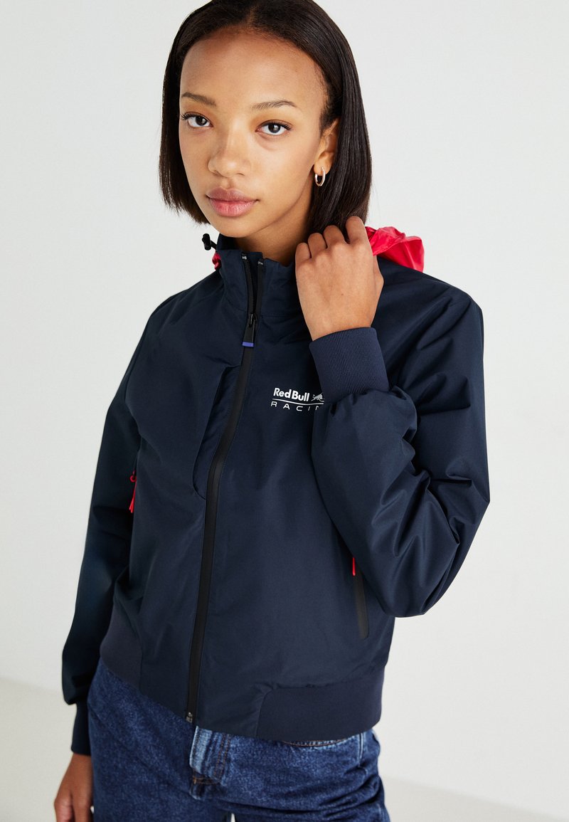 Chaqueta cortavientos de color marino con cuello alto, cremallera negra, cordón rosa y puños elásticos. Logo de "Red Bull Racing" en el pecho.