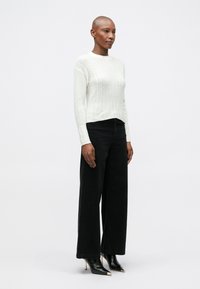 Sweater branco de malha canelada com mangas longas, combinado com jeans de perna larga pretos de cintura alta e sapatos de salto pretos pontiagudos com detalhes em dourado.