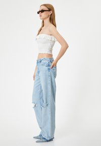 Top sin tirantes blanco con smock, vaqueros de pierna ancha azul claro con desgastes, y zapatos planos azules de punta. Gafas de sol y pendientes de aro completan el look.