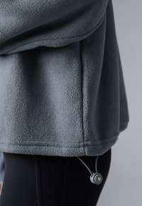 Pullover in pile grigio con finitura testurizzata, dotato di cordino e toggle all'orlo, abbinato a leggings neri.