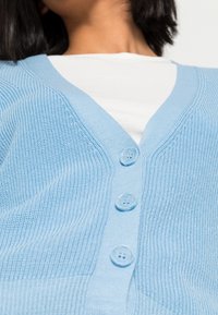 Ljusblå stickad cardigan med V-ringning, prydd med tre knappar framtill och ribbad textur, lagd över en vit skjorta.
