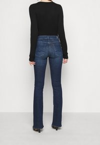 Jean évasé en denim de lavage foncé avec une taille mi-haute ; comporte des poches arrière avec des détails cousus et une texture de denim lisse.