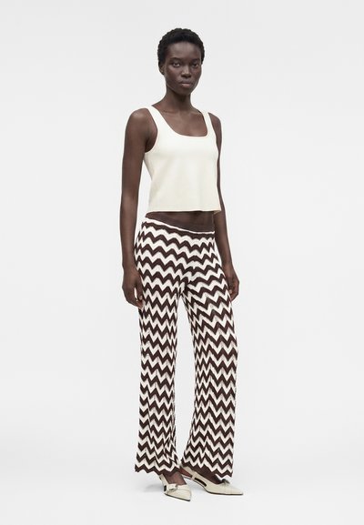 Γυναίκα που στέκεται, φοράει λευκό αμάνικο τοπ, παντελόνι φαρδιά με ασπρόμαυρα ζιγκ-ζαγκ σχέδια και λευκά μυτερά παπούτσια.