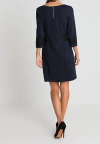 Mörkblå klänning med trekvartsärmar, med svarta spetsdetaljer vid ärmsluten och sidan, samt dragkedja bak. Slät textur.