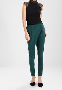 Top negro sin mangas con escote de encaje, combinado con pantalones verde ajustados con pliegues y cinturilla elástica. Zapatos de tacón negros.