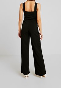 Combinaison noire avec un design à jambes larges, bretelles épaisses et un corsage ajusté. Texture lisse, style minimaliste et ourlet à la cheville.