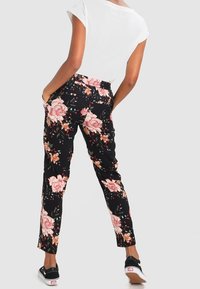 Person som bär svarta byxor med rosa blommönster, vit kortärmad topp och svarta slip-on skor, står med händerna i fickorna och vänder sig bort.