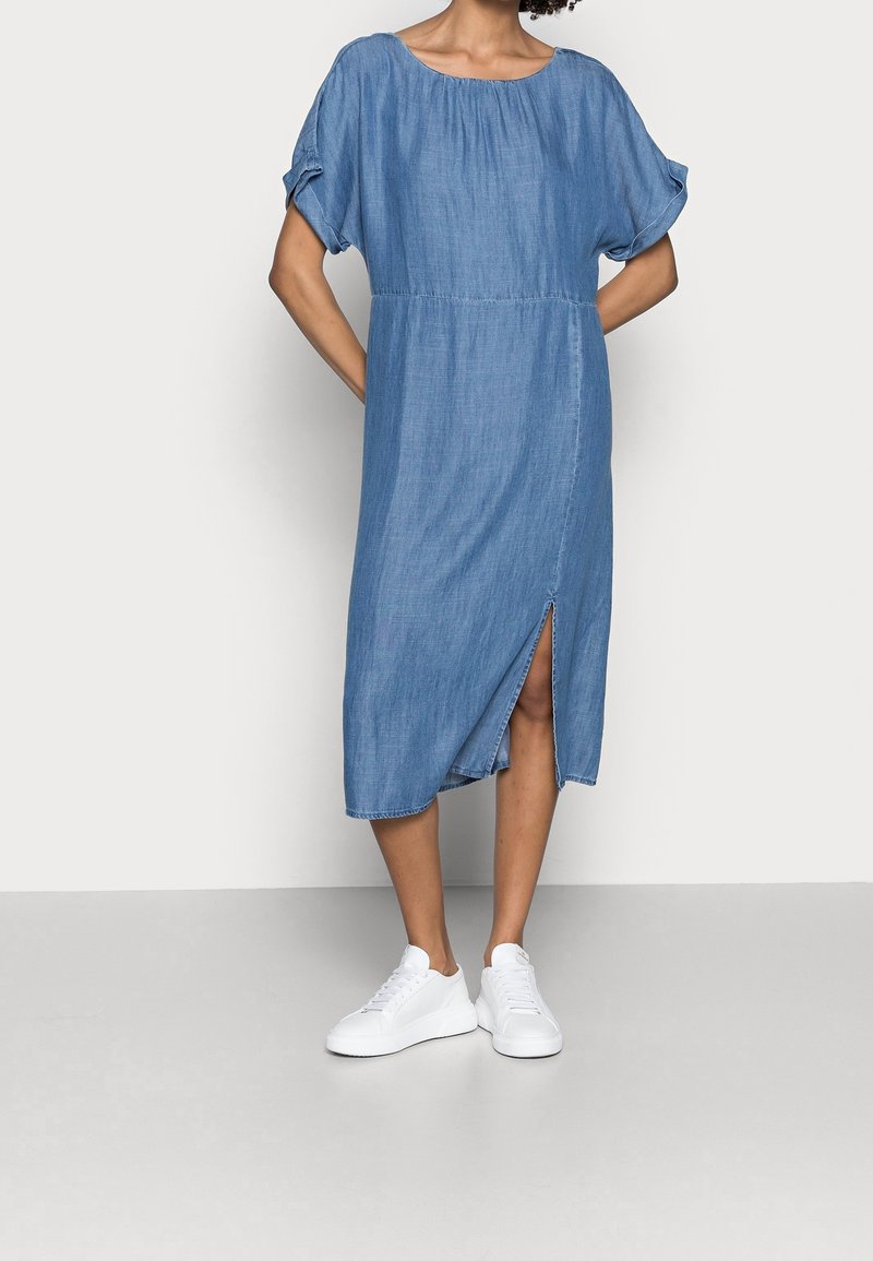 Robe midi en denim, bleu clair, coupe ample avec des manches courtes bouffantes et un col rond. Détail de fente sur le côté, portée avec des baskets blanches.