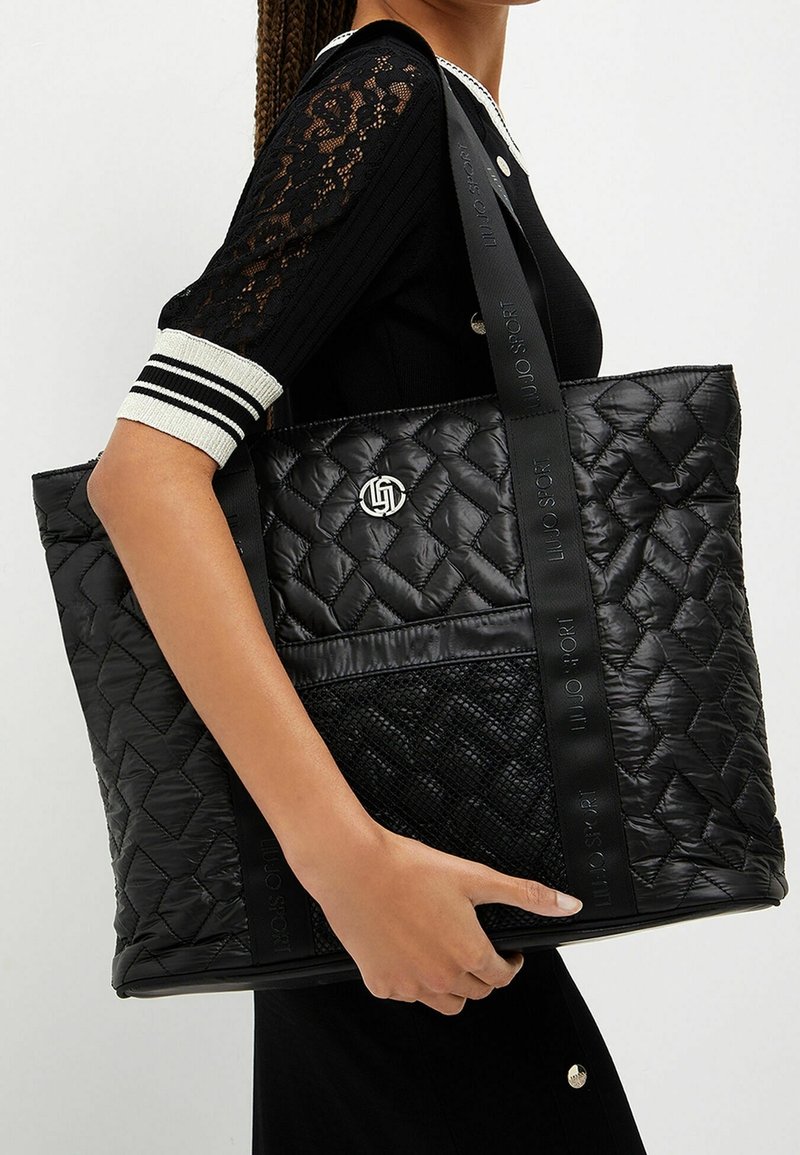 Femme portant un haut noir à manches en dentelle, portant un grand sac cabas noir matelassé avec des anses marquées et un badge logo argenté.