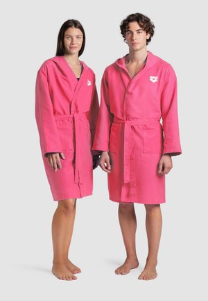 Un uomo e una donna sono in piedi a piedi nudi, indossando accappatoi rosa fino al ginocchio con cinture e loghi "arena" sul petto, su uno sfondo semplice.