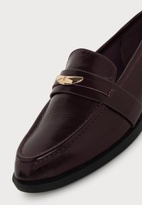 Mocassin en cuir bordeaux foncé avec une finition brillante, semelle noire et petite boucle décorative dorée sur la bride supérieure.