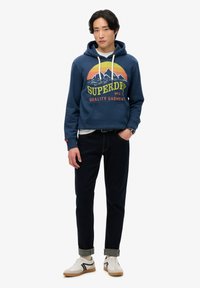 Sudadera azul marino con gráfico de montaña y texto "SUPERDRY", combinada con jeans oscuros y zapatillas blancas con detalles en negro.