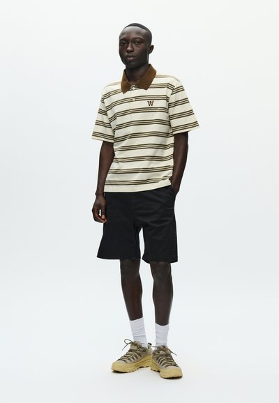 Polo rayé crème et marron, manches courtes, avec col et logo. Associé à un short noir et des baskets beiges.