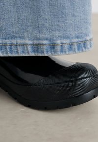Black rubber shoe with a textured finish and smooth toe, paired with blue denim pant cuffs resting above the shoe.