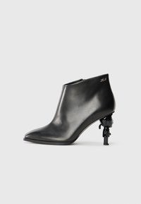 Black leather ankle boot with pointed toe, glossy finish, and distinctive sculptural heel featuring a figurine accent. Side zip closure.