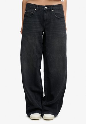 Løstsiddende sorte denimjeans båret med hvide sneakers, synlige fra taljen til fødderne, mod en ensfarvet baggrund.
