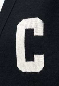 Witte gebreide letter "C" op zwart gebreide stof, met zichtbare textuur en stikseldetails van het kledingstuk.