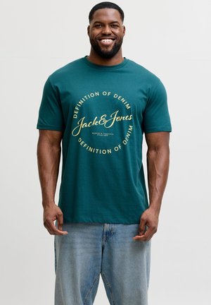 Smiling man wearing teal Jack & Jones t-shirt with "Definition of Denim" text, paired with light blue jeans, standing against white background.