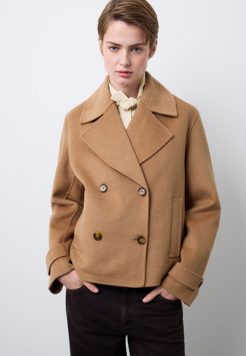 Braune, zweireihige Wolljacke mit großen Knöpfen, Reverskragen und Seitentaschen; getragen über einer cremefarbenen Bluse mit glatter Textur.