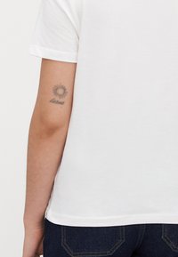 Asmens kairė ranka su maža saulės tatuiruote ir kursyvine užrašu "karma", matoma po trumpa balta rankove ir virš tamsiai džinsų.