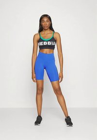 Čierny crop top s bielym nápisom „NEBBIA“ a zelenými akcentmi, v kombinácii s modrými cyklistickými šortkami. Modelka má na nohách čierne športové topánky.