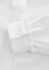 Weißes Hemd mit Knöpfen in einem strukturierten Stoff, Rüschendetail an den Manschetten und normalen Knöpfen. Kragen und Manschetten sind ordentlich gebügelt.