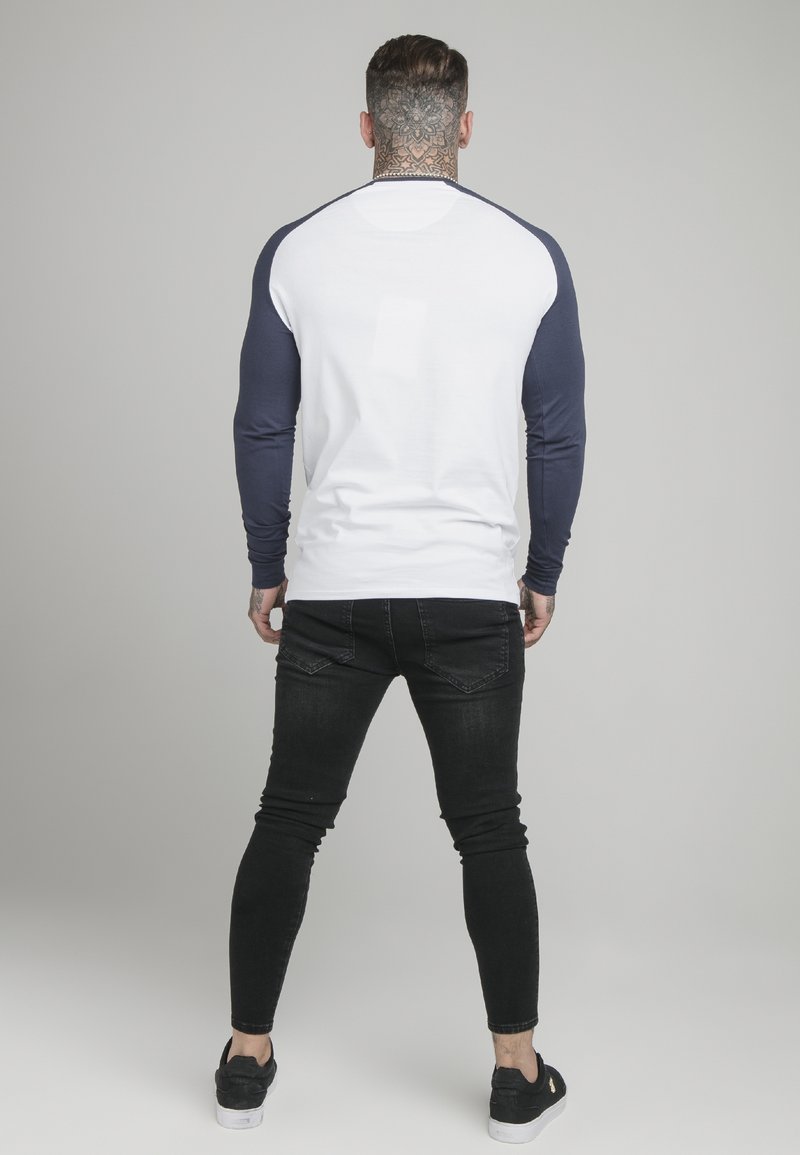 Mann mit floralem Nackentattoo, der ein weißes und navyblaues Langarmhemd, schwarze enge Jeans und schwarze Sneakers trägt, steht vor einem schlichten grauen Hintergrund.