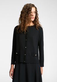 Svart kardigan av mjukt material, med rund hals, knapplist och dekorativa framfickor med accenter.