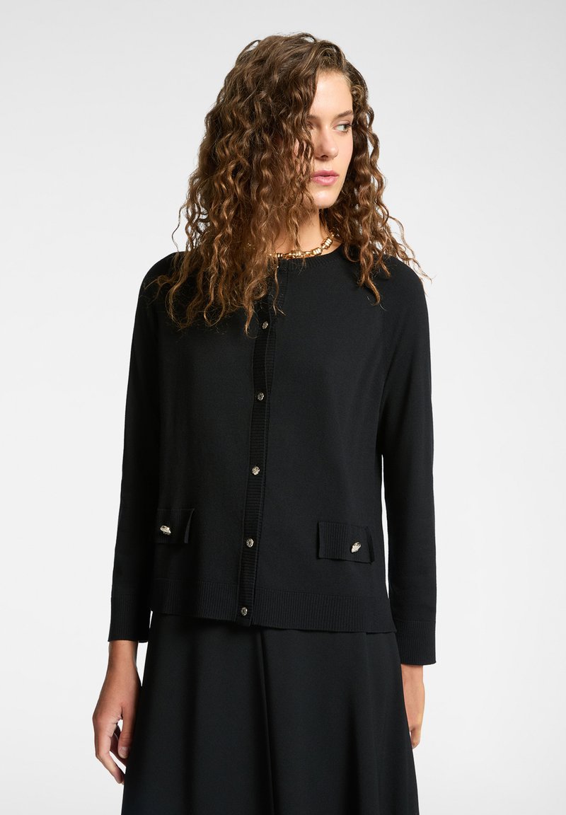 Svart kardigan av mjukt material, med rund hals, knapplist och dekorativa framfickor med accenter.